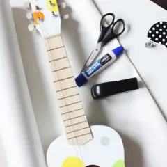 Przygotowania do wysyłki gitary: biała gitara, nożyczki, klej, zszywacz i ozdobna kokardka.