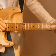 Dłoń grająca na gitarze elektrycznej, z widocznym gryfem i strunami. Delikatne światło podkreśla drewno i **ponticello** dźwięku.