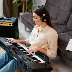 Kobieta w słuchawkach gra na keyboardzie japońskiej marki. Obok stoi wzmacniacz i gitara.