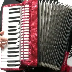 Ręce grają na czerwonym akordeonie. Widoczna klawiatura i miech, ukazujące budowę instrumentu.