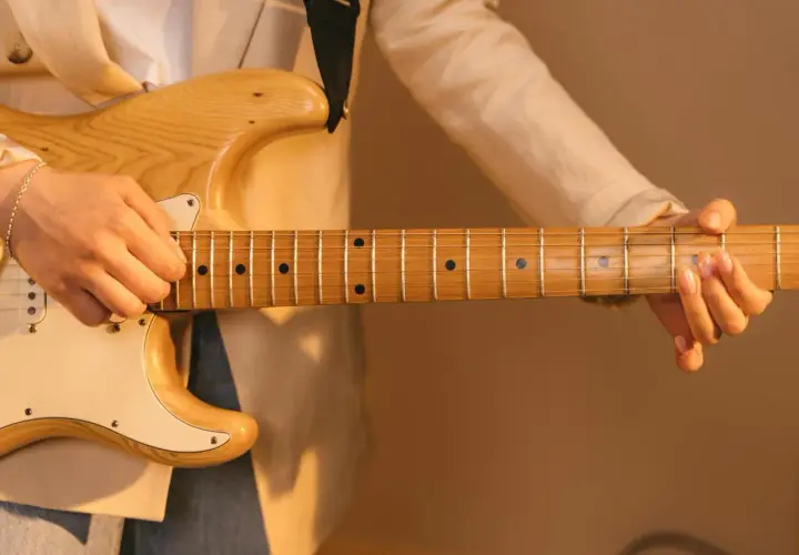 Dłoń grająca na gitarze elektrycznej, z widocznym gryfem i strunami. Delikatne światło podkreśla drewno i **ponticello** dźwięku.