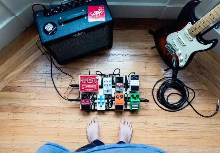 Stojący na drewnianej podłodze pedalboard z efektami gitarowymi, gotowy do gry. W tle wzmacniacz i gitara.