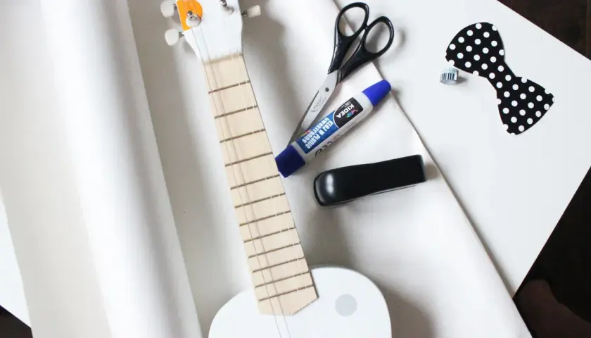 Przygotowania do wysyłki gitary: biała gitara, nożyczki, klej, zszywacz i ozdobna kokardka.