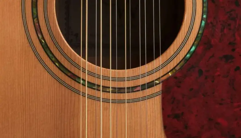 Zbliżenie na gitarę 12 strunową, widoczne dwanaście strun, rozetę z masy perłowej i czerwony pickguard. Strojenie gitary 12 strunowej to wyzwanie.