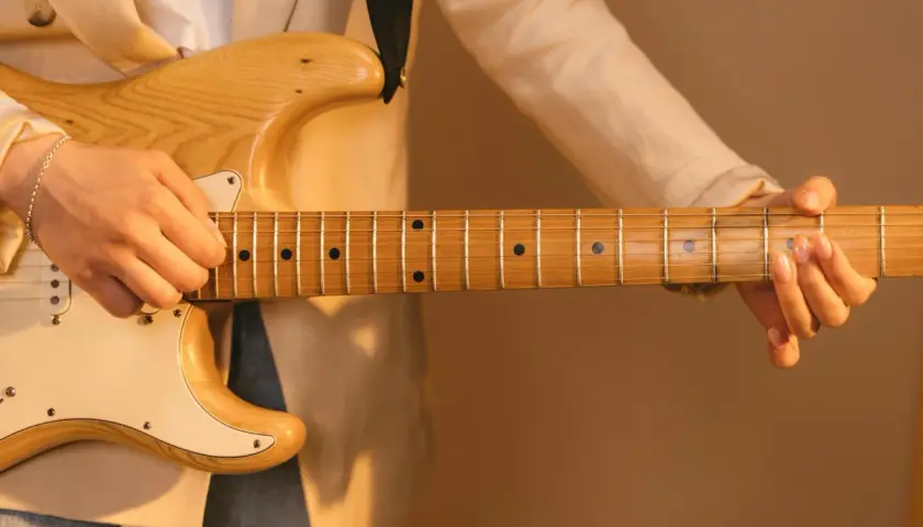 Dłoń grająca na gitarze elektrycznej, z widocznym gryfem i strunami. Delikatne światło podkreśla drewno i **ponticello** dźwięku.