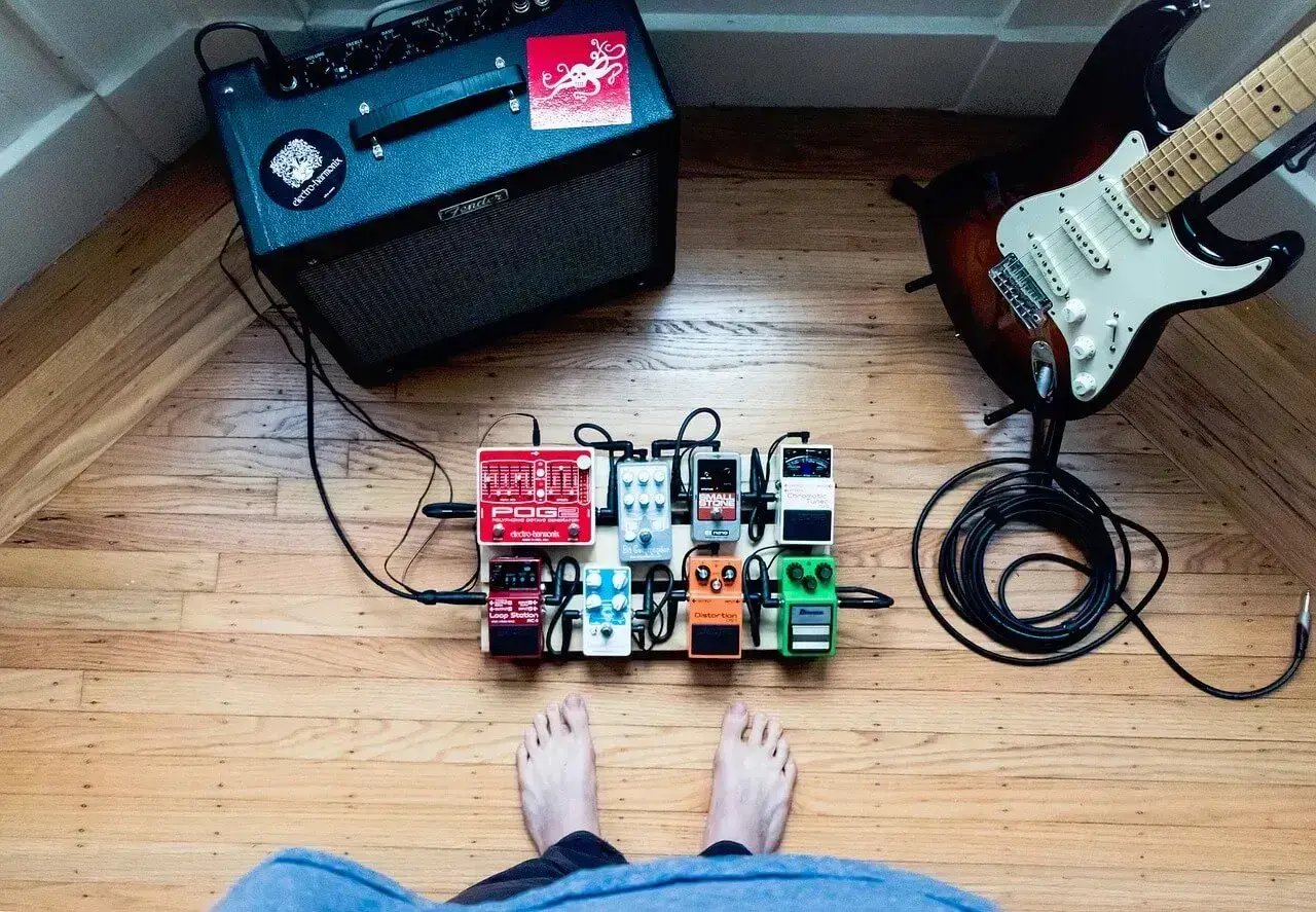 Stojący na drewnianej podłodze pedalboard z efektami gitarowymi, gotowy do gry. W tle wzmacniacz i gitara.