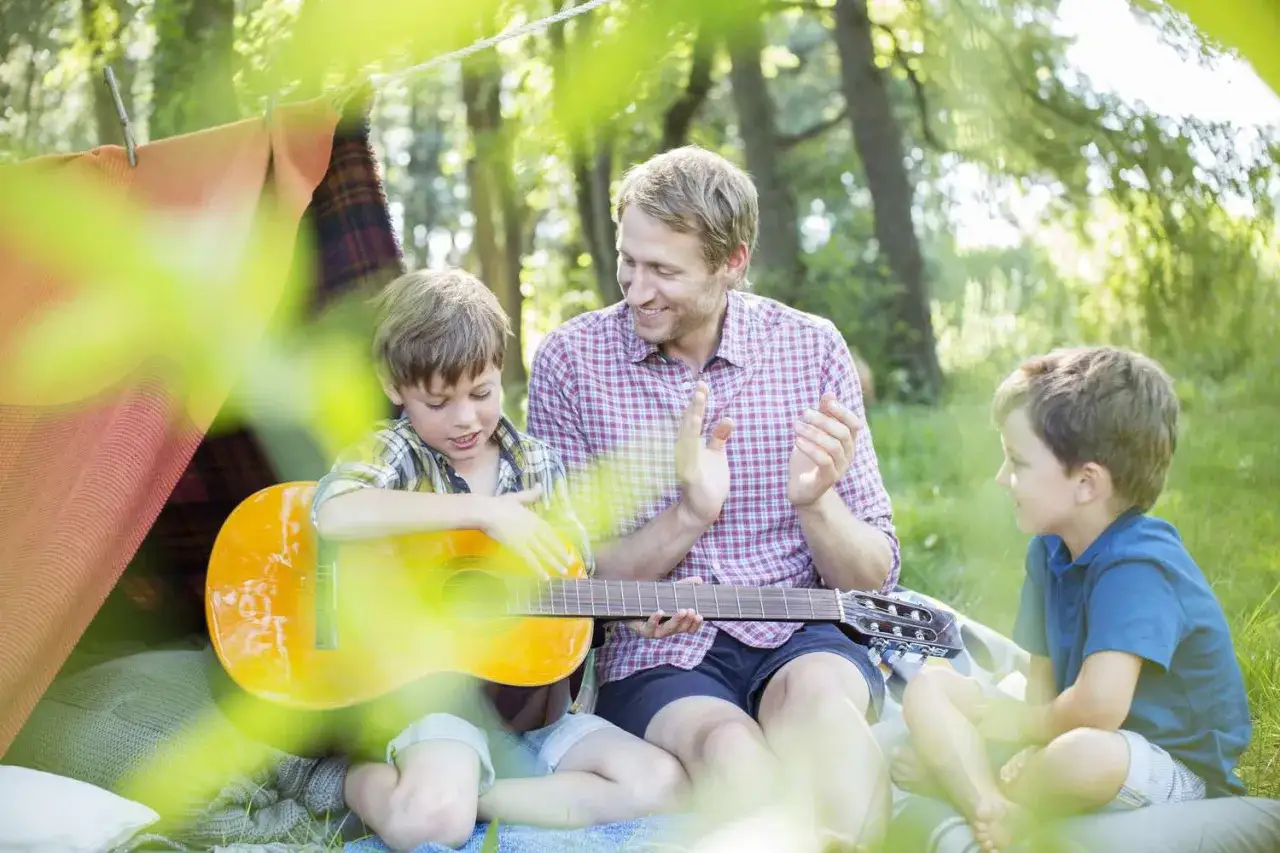 Ojciec z synami na pikniku, chłopiec gra na gitarze, a tata klaszcze. W tle namiot i drzewa. Wszyscy uczą się chwyty alleluja.