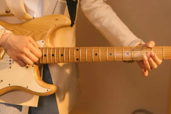Dłoń grająca na gitarze elektrycznej, z widocznym gryfem i strunami. Delikatne światło podkreśla drewno i **ponticello** dźwięku.